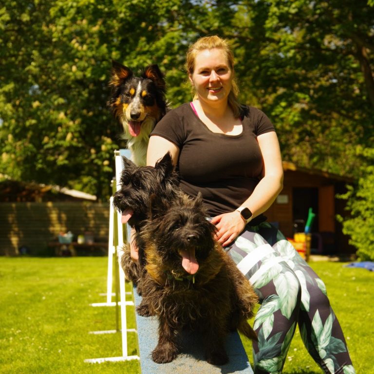 Andrea Knoll, Tierphysiotherapeutin Andrea Knoll, die Tierphysiotherapeutin, sitzt auf einer Agility-Hindernisbahn mit drei Hunden im Freien.